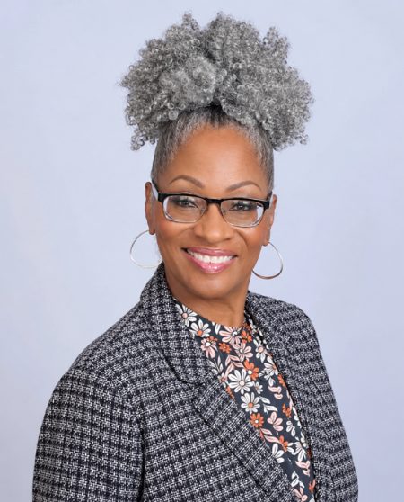 Professional headshot of a woman with gray hair in a high puff style, wearing glasses, hoop earrings, and a patterned cardigan over a floral top