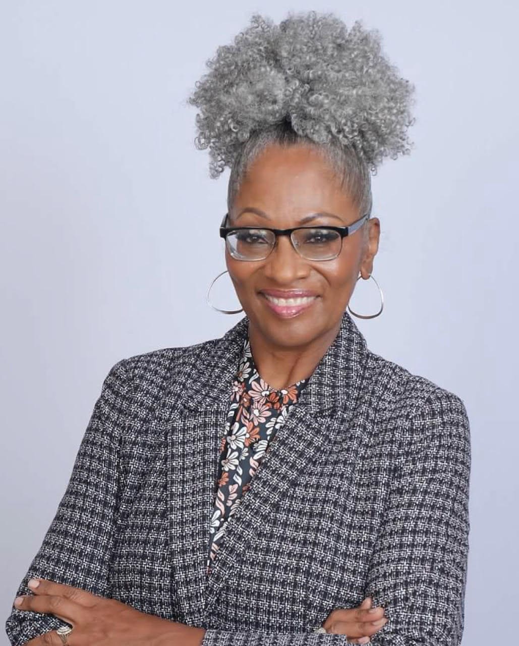 Professional headshot of a woman with gray hair in a top bun, wearing glasses and a dark textured blazer